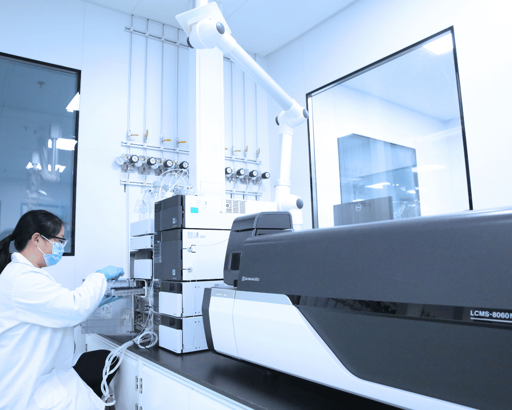 Apeloa woman scientist in white lab coat in front of an LCMS instrument in analytical development lab.