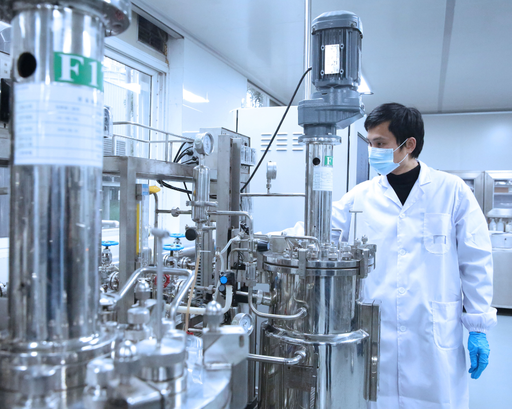 Analyst in white lab coat, wearing mask and gloves, in front of stainless steel bio-production fermentation reactors.