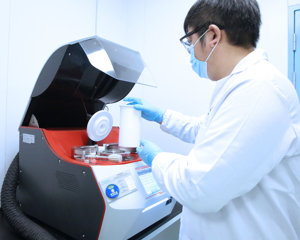 Male analyst in a white h=lab coat standing in front of a red and black lab instrument performing and analytical test.