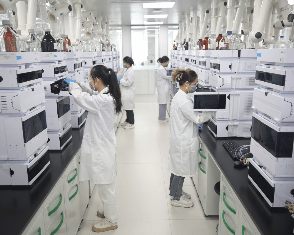 Apeloa CDMO lab containing more than 10 white HPLC systems on benchtops, operated by two scientists in white lab coats.