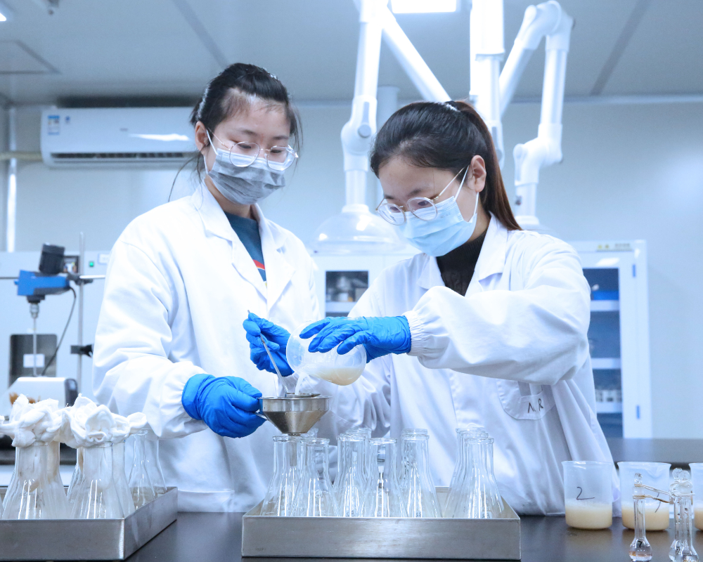 Two lab analysts in white lab coats, and blue masks and gloves sample fermenation samples in the bio-production laboratory.