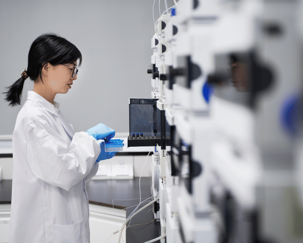 Woman scientist in white lab coat standing beside a row of white HPLC analytical instruments.