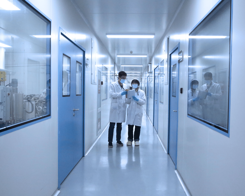 Two scientists in white labcoats walking down white pharmaceutical facility hallway.